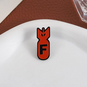 Detail of red bomb-shaped enamel pin with black letter F on a white ceramic plate
