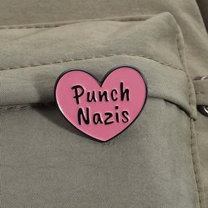Close-up of a pink heart enamel pin with black text on an olive green fabric