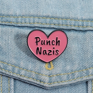 Close-up of a pink heart-shaped enamel pin with Punch Nazis text on blue denim fabric