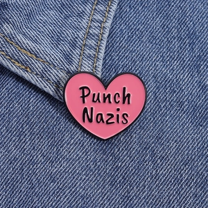 Close-up of a pink heart-shaped enamel pin with Punch Nazis text on denim fabric