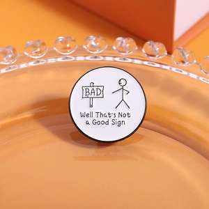 Close-up of a round enamel pin featuring a stick figure next to a BAD sign and witty text