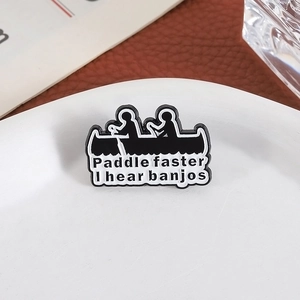 Black enamel canoe brooch with Paddle faster I hear banjos text on a white plate