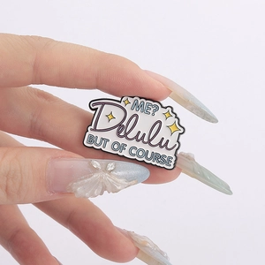 Vue détaillée du badge en émail Delulu tenu à la main sur un fond blanc épuré