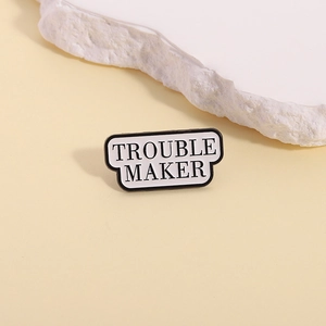 Enamel brooch pin featuring Trouble Maker text in black and white on a stony background