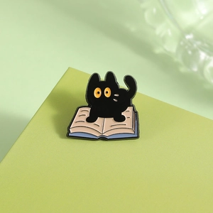 Close-up of a cute black cat sitting on an open book enamel pin on a green background