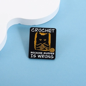 Detail of square black enamel pin featuring a yellow cat crocheting with white text
