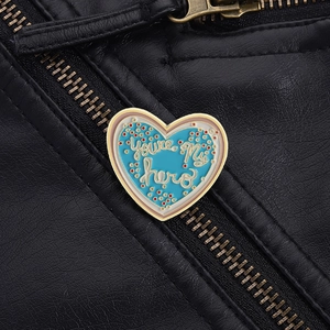 Dettaglio della spilla a cuore smaltata blu con testo You're My Hero su giacca nera
