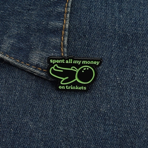 Detail of green enamel pin on denim featuring spent all my money on trinkets text