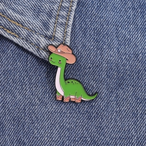 Close-up detail of green dinosaur enamel pin wearing a cowboy hat on denim fabric