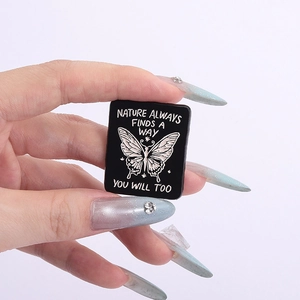 Hand holding black enamel brooch featuring a white butterfly and inspirational quote