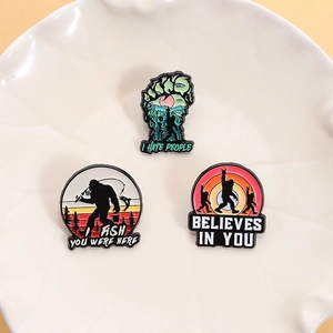 Three Big Foot enamel pins with forest and fishing themes displayed on a white plate