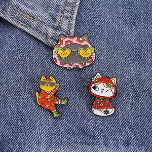 Three colorful cat enamel pins with sunglasses and floral patterns on blue denim fabric