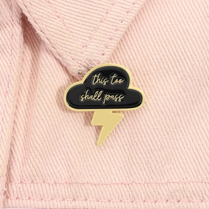 Detail of storm cloud enamel pin with this too shall pass text on pink denim fabric