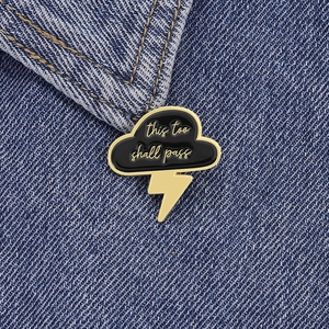Minimalist cloud and lightning enamel pin with 'this too shall pass' quote on denim fabric