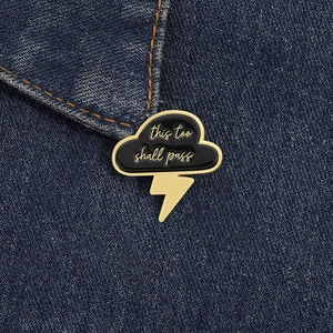Macro detail of cloud and lightning enamel pin with 'this too shall pass' text on denim