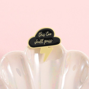 Detail of cloud and lightning enamel pin with This Too Shall Pass engraving on shell