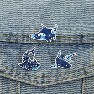 Detail of three ocean-themed enamel shark pins with wave patterns on a blue denim jacket
