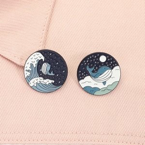 Detail view of two whale enamel pins with sea waves and moon patterns on pink fabric