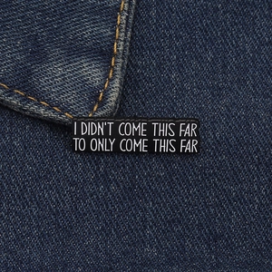 Detailed close-up of rectangular motivational enamel pin on dark denim fabric background
