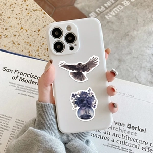 White iPhone case with artistic dark crow and floral rose illustrations in a lifestyle setting