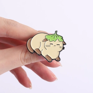 Detail shot of a cute beige cat enamel pin with a green leaf on its head held in hand