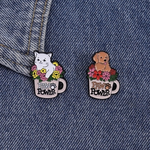 Detail of enamel pins featuring a cat and dog in flower coffee cups on denim