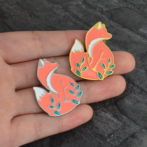 Détail de deux broches renard en émail orange avec motifs floraux tenues en main