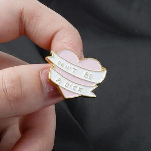 Pink heart enamel pin with white ribbon and Don't Be A Dick text on gray fabric
