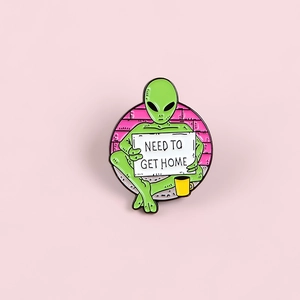 Detailed close-up of green alien enamel pin holding a 'NEED TO GET HOME' sign on pink background