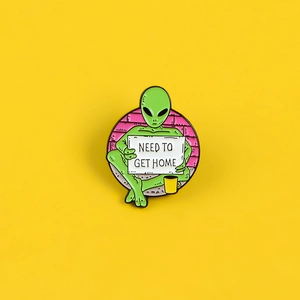 Detail of green alien enamel pin with NEED TO GET HOME sign on yellow background