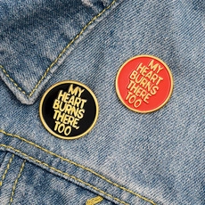 Detail of round black and red enamel pins with gold My Heart Burns There Too text on denim
