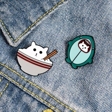 Detail of enamel cat pins featuring white cat in rice bowl and shark costume on denim fabric