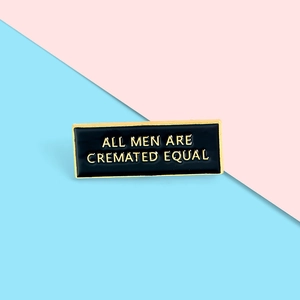 Macro detail of black enamel pin with gold lettering All Men Are Cremated Equal