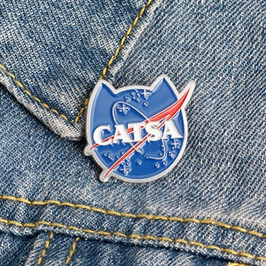 Blue cat-shaped enamel pin with NASA parody CATSA text on denim jacket background