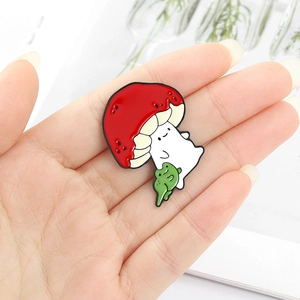 Detailed close-up of a cartoon red mushroom and small green frog enamel pin on a hand