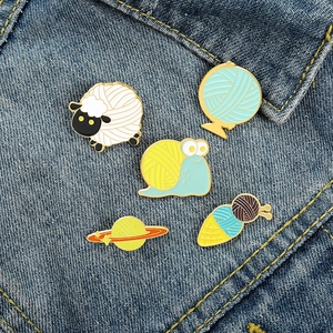 Close-up of five creative knitting-themed enamel pins displayed on blue denim fabric
