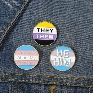 Drei Emaille-Pins mit Pronomen wie They/Them und He/Him auf blauem Jeansstoff Hintergrund