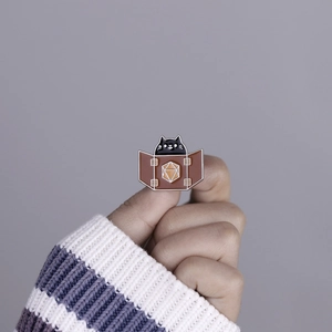 Close-up of a cute black cat in a box enamel pin held against a purple sweater