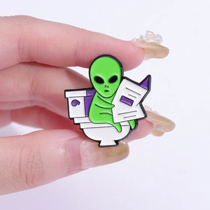 Detailed close-up of a funny green ET alien enamel pin sitting on a toilet reading a newspaper