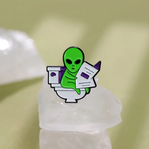 Close-up of a funny green ET alien enamel pin sitting on a toilet reading a newspaper