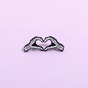 Broche en émail représentant des mains de squelette formant un cœur en noir et blanc