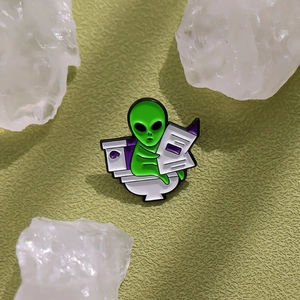 Funny green alien enamel pin sitting on a toilet reading a newspaper detail view