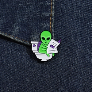 Close-up of funny green alien sitting on toilet reading newspaper enamel pin on denim