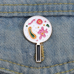 Macro di una spilla smaltata a lente d'ingrandimento dorata con batteri colorati su denim