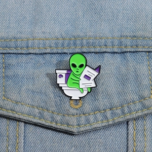 Funny green alien enamel pin on blue denim jacket reading on a white toilet