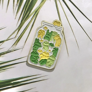 Close-up of creative frog jar enamel pin with green cartoon frogs and gold metal edges