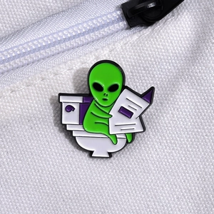 Detail shot of a funny green alien enamel pin sitting on a white toilet reading a newspaper