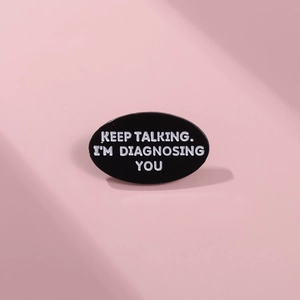 Détail d'une broche ovale en émail noir avec la phrase d'humour Keep Talking I Am Diagnosing You