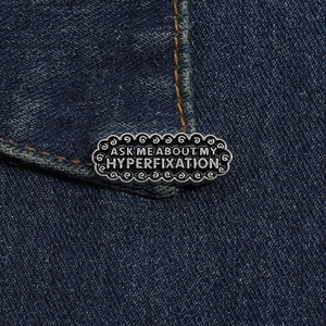 Detail of ASK ME ABOUT MY HYPERFIXATION enamel pin on a blue denim pocket edge