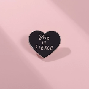 Macro detail of a black heart enamel pin featuring the silver She IS FIERCE lettering